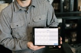 A person is holding a tablet displaying a point-of-sale system. The person is wearing a gray button-up shirt and stands in a room with dark walls and shelves in the background. The tablet screen shows various products and prices, indicating a business environment.