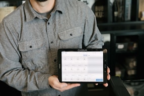 A person is holding a tablet displaying a point-of-sale system. The person is wearing a gray button-up shirt and stands in a room with dark walls and shelves in the background. The tablet screen shows various products and prices, indicating a business environment.