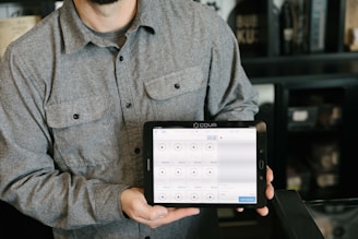 A person is holding a tablet displaying a point-of-sale system. The person is wearing a gray button-up shirt and stands in a room with dark walls and shelves in the background. The tablet screen shows various products and prices, indicating a business environment.