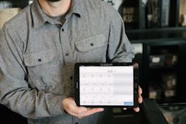 A person is holding a tablet displaying a point-of-sale system. The person is wearing a gray button-up shirt and stands in a room with dark walls and shelves in the background. The tablet screen shows various products and prices, indicating a business environment.
