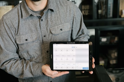 A person is holding a tablet displaying a point-of-sale system. The person is wearing a gray button-up shirt and stands in a room with dark walls and shelves in the background. The tablet screen shows various products and prices, indicating a business environment.