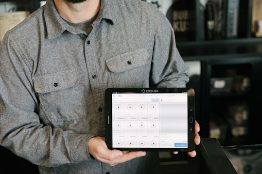 A person is holding a tablet displaying a point-of-sale system. The person is wearing a gray button-up shirt and stands in a room with dark walls and shelves in the background. The tablet screen shows various products and prices, indicating a business environment.
