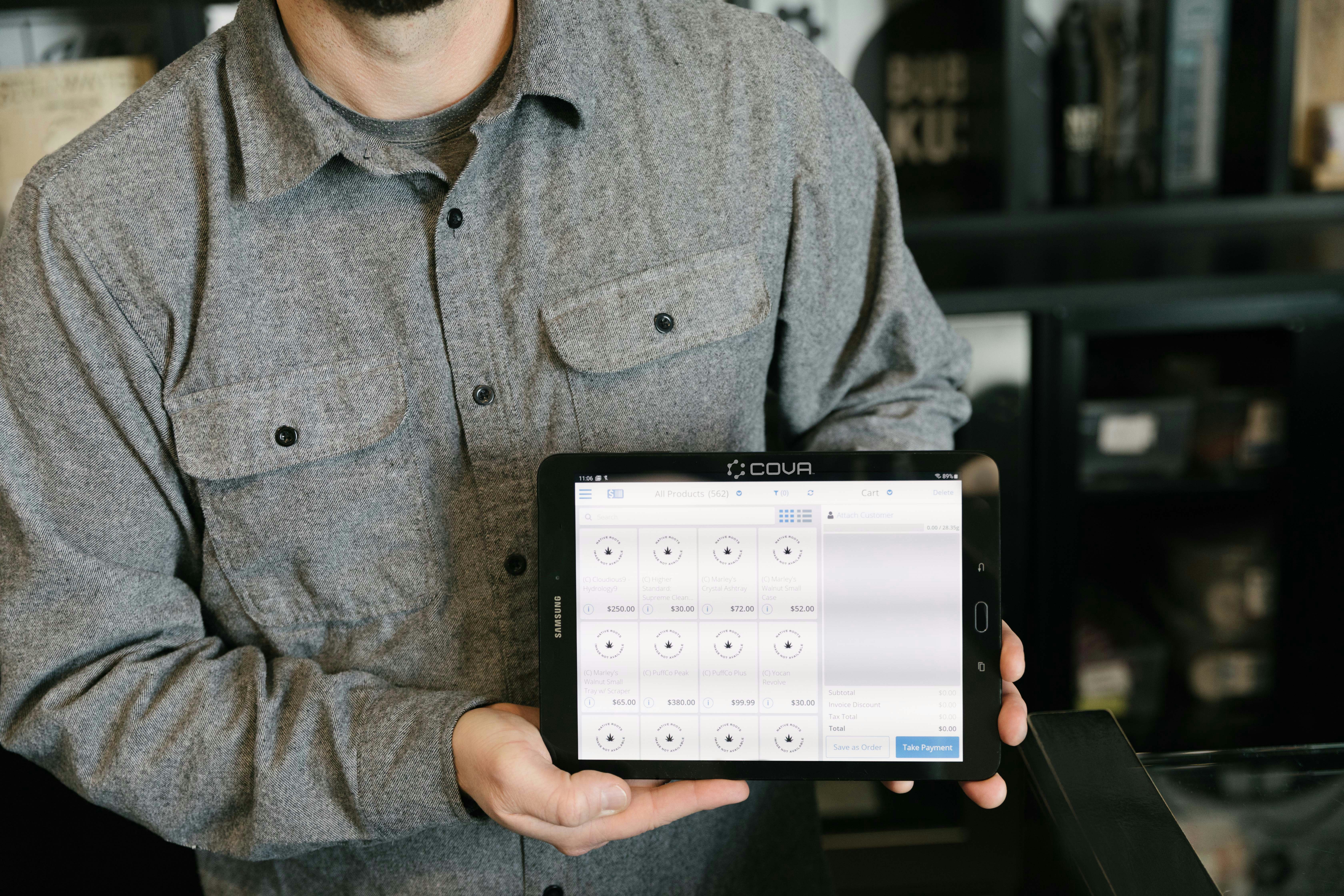 A male budtender in a dispensary displays the strains or cultivars available for cannabis consumers to purchase on a lit tablet. The product information is arranged and displayed using Cova's cannabis point of sale software.