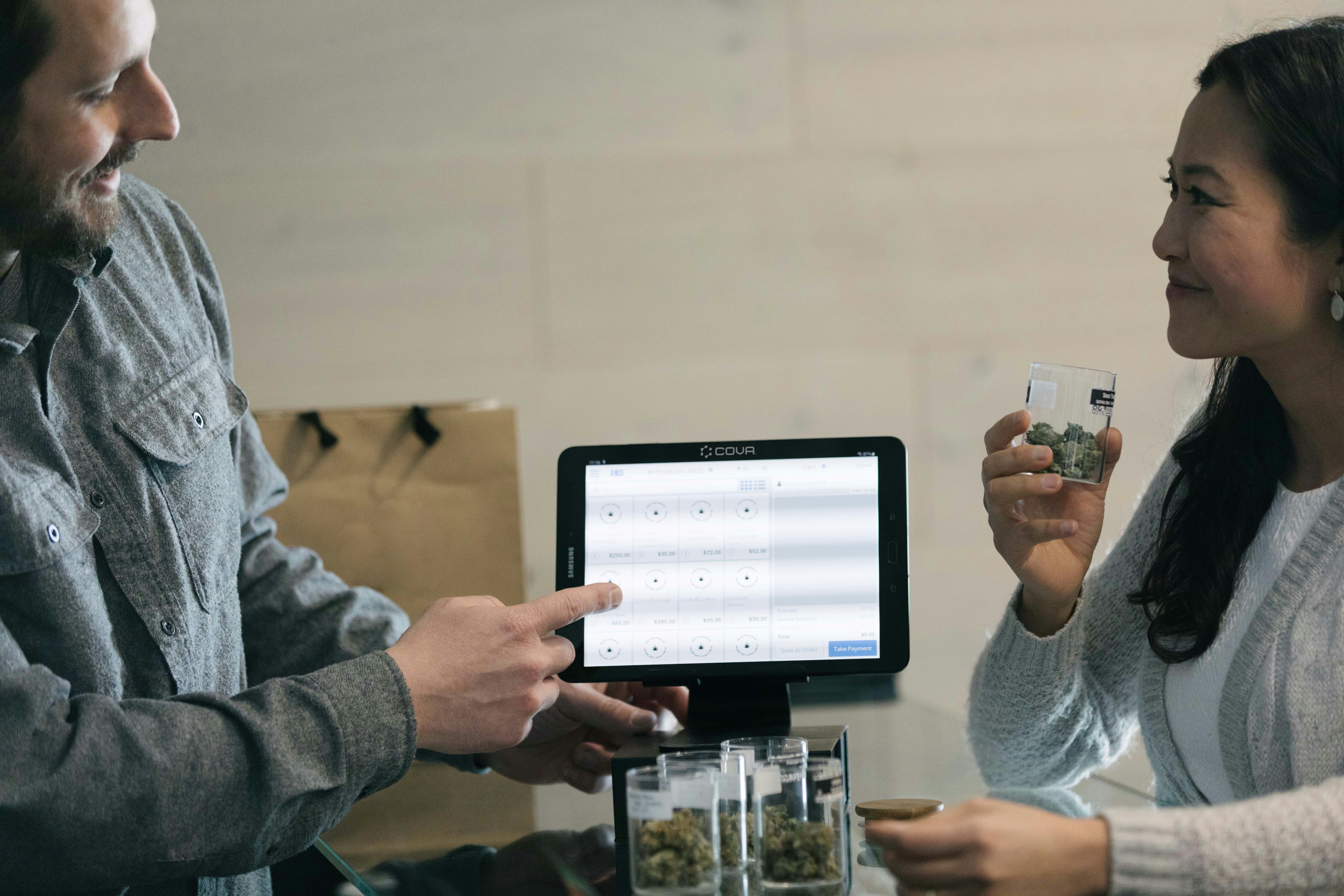 A male budtender and female cannabis customer discuss the merits of various cultivars in a dispensary. The budtender points to a lit tablet that is between them and displays Cova's point of sale cannabis software while the customer considers the product.