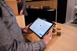 A person holding a tablet device with a digital interface displayed on the screen, possibly related to a point-of-sale or inventory system. The person is standing at a wooden counter, on which there are two jars containing dried green plant material. Another person is in the background, partially visible, standing next to a cardboard box.