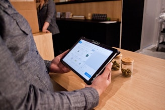 A person holding a tablet device with a digital interface displayed on the screen, possibly related to a point-of-sale or inventory system. The person is standing at a wooden counter, on which there are two jars containing dried green plant material. Another person is in the background, partially visible, standing next to a cardboard box.