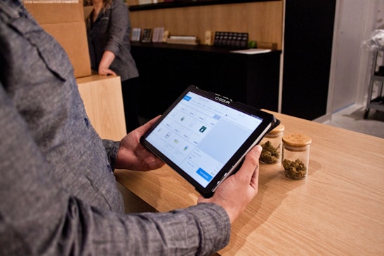 A person holding a tablet device with a digital interface displayed on the screen, possibly related to a point-of-sale or inventory system. The person is standing at a wooden counter, on which there are two jars containing dried green plant material. Another person is in the background, partially visible, standing next to a cardboard box.