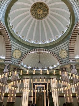 A beautifully decorated mosque interior featuring an ornate dome with intricate Islamic geometric patterns and calligraphy in gold and green. A large circular chandelier with numerous white lanterns hangs below the dome, illuminating the space. The space is supported by large columns with decorative designs.