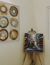 A wooden easel holds a vibrant and colorful abstract painting, depicting a lively street scene with figures and vehicles. Adjacent on the wall, a framed artwork features six concentric circles in earthy tones, arranged in a grid pattern. The setting is an interior space with neutral wall colors, softly lit and featuring a window to the right.