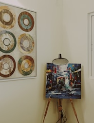 A wooden easel holds a vibrant and colorful abstract painting, depicting a lively street scene with figures and vehicles. Adjacent on the wall, a framed artwork features six concentric circles in earthy tones, arranged in a grid pattern. The setting is an interior space with neutral wall colors, softly lit and featuring a window to the right.