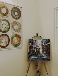 A wooden easel holds a vibrant and colorful abstract painting, depicting a lively street scene with figures and vehicles. Adjacent on the wall, a framed artwork features six concentric circles in earthy tones, arranged in a grid pattern. The setting is an interior space with neutral wall colors, softly lit and featuring a window to the right.
