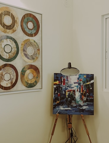 A wooden easel holds a vibrant and colorful abstract painting, depicting a lively street scene with figures and vehicles. Adjacent on the wall, a framed artwork features six concentric circles in earthy tones, arranged in a grid pattern. The setting is an interior space with neutral wall colors, softly lit and featuring a window to the right.