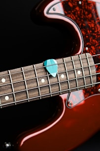 A close-up of a guitar focusing on its fretboard and a teal guitar pick placed on the strings. The body of the guitar features a rich amber color with a glossy finish and a tortoiseshell pickguard. The strings and frets are clearly visible, highlighting the intricate craftsmanship of the instrument.