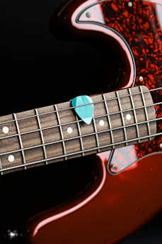 A close-up of a guitar focusing on its fretboard and a teal guitar pick placed on the strings. The body of the guitar features a rich amber color with a glossy finish and a tortoiseshell pickguard. The strings and frets are clearly visible, highlighting the intricate craftsmanship of the instrument.