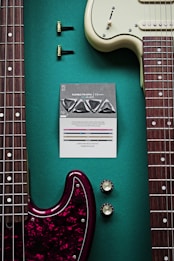 A close-up view of two electric guitars placed parallel to each other. One guitar has a burgundy pickguard and the other has a cream-colored pickguard. A set of guitar picks is placed in the center, along with tuning pegs and control knobs. The background is a teal color, providing a vibrant contrast to the various guitar elements.