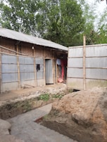 A rural scene featuring a simple structure made with metal sheets and bamboo. An earthen trench or excavation is visible in the foreground, indicating construction work. Clothes hang on the side of the building, and lush green trees surround the area.
