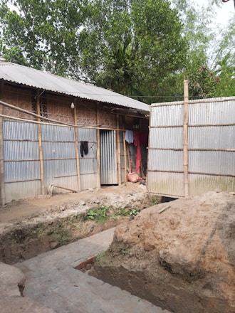 A rural scene featuring a simple structure made with metal sheets and bamboo. An earthen trench or excavation is visible in the foreground, indicating construction work. Clothes hang on the side of the building, and lush green trees surround the area.