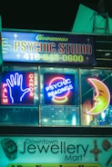 Neon signs for a psychic studio and jewelry mart are displayed at night. The psychic studio sign features bright neon lights with the words 'Palm Cards' and 'Psychic Readings', along with iconic images like a hand and a crescent moon. Below it, a jewelry shop sign is visible.
