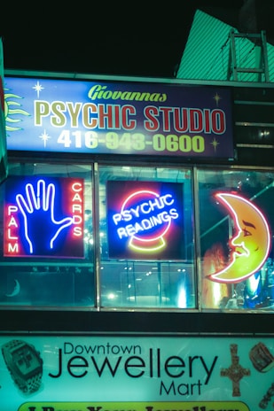 Neon signs for a psychic studio and jewelry mart are displayed at night. The psychic studio sign features bright neon lights with the words 'Palm Cards' and 'Psychic Readings', along with iconic images like a hand and a crescent moon. Below it, a jewelry shop sign is visible.