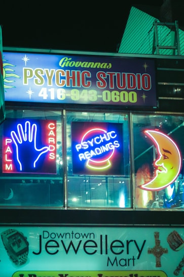 Neon signs for a psychic studio and jewelry mart are displayed at night. The psychic studio sign features bright neon lights with the words 'Palm Cards' and 'Psychic Readings', along with iconic images like a hand and a crescent moon. Below it, a jewelry shop sign is visible.