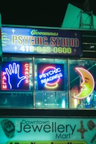 Neon signs for a psychic studio and jewelry mart are displayed at night. The psychic studio sign features bright neon lights with the words 'Palm Cards' and 'Psychic Readings', along with iconic images like a hand and a crescent moon. Below it, a jewelry shop sign is visible.