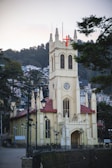 A tall, Gothic-style church building stands prominently with a red-roofed tower and stained-glass windows. The structure is surrounded by iron lamp posts and fencing, set against a backdrop of hills and a clustered residential area.