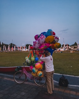 A vendor stands beside a bicycle filled with colorful balloons and toys. The setting appears to be an open park with flags in the background. The sky is clear with a hint of blue, creating a serene atmosphere.