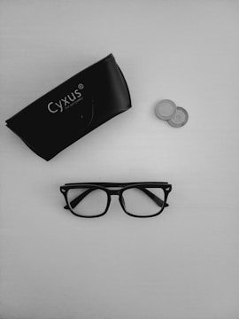 A pair of black-rimmed glasses is positioned below a black glasses case labeled 'Cyxus', with a couple of coins nearby on a light surface.