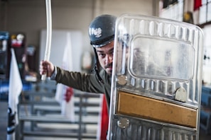 A person wearing a helmet and holding a baton stands behind a transparent riot shield. The setting appears to be indoors with benches and flags visible in the background.
