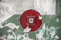 A red fire alarm button is set against a weathered wall with peeling paint, featuring shades of green and white.