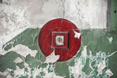 A red fire alarm button is set against a weathered wall with peeling paint, featuring shades of green and white.