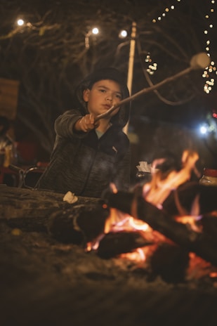 Children roasting marshmallows around a campfire beside a cabin at dusk.