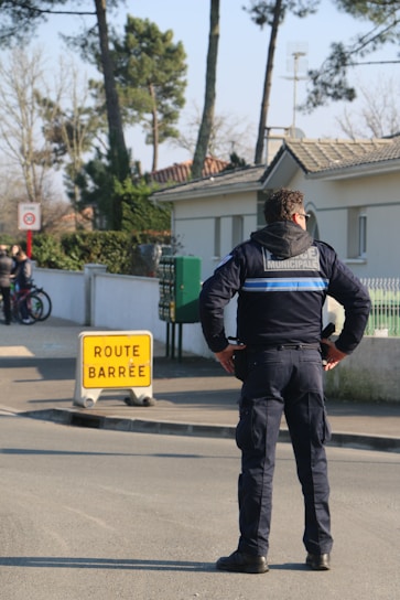 A municipal officer is standing on a road with hands on hips, facing a 'route barrée' (road closed) sign. The environment includes a residential area with trees and houses nearby. In the background, several people are seen, some with bicycles.