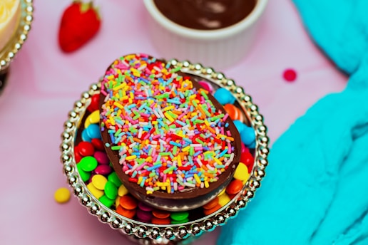 A chocolate egg topped with bright, colorful sprinkles sits on a silver dish filled with colorful candy-coated chocolates. In the background, there is a strawberry, a small bowl of chocolate sauce, and a blue fabric.