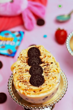 A dessert topped with cream and a row of chocolate cookies is placed on a decorative silver stand. The background features a playful, pink plush toy, a blue package with branding, a whole strawberry, a spoon, and chocolate cookie pieces scattered around.