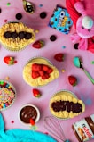 An assortment of colorful desserts is arranged on a pink surface. The selection includes cupcakes topped with mini Oreo cookies, vibrant rainbow sprinkles, and fresh strawberries. Accompanying the treats are small candy-coated chocolates scattered around, a jar of Nutella, and a small bowl filled with chocolate sauce. A plush pink stuffed animal, possibly a toy, and a metallic whisk add a playful touch to the scene.