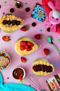 An assortment of colorful desserts is arranged on a pink surface. The selection includes cupcakes topped with mini Oreo cookies, vibrant rainbow sprinkles, and fresh strawberries. Accompanying the treats are small candy-coated chocolates scattered around, a jar of Nutella, and a small bowl filled with chocolate sauce. A plush pink stuffed animal, possibly a toy, and a metallic whisk add a playful touch to the scene.
