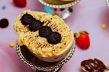 A dessert topped with creamy frosting and three chocolate cookies arranged in a row. It is placed on a decorative silver stand, surrounded by a background that includes a strawberry, scattered chocolate pieces, and colorful candy on a pink surface.