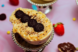 A dessert topped with creamy frosting and three chocolate cookies arranged in a row. It is placed on a decorative silver stand, surrounded by a background that includes a strawberry, scattered chocolate pieces, and colorful candy on a pink surface.