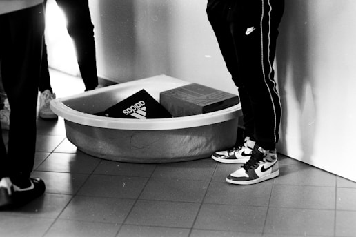 A black and white photograph showing a large plastic container placed on a tiled floor. Inside the container, there are two shoeboxes, one from Adidas and another possibly from Nike. Surrounding the container are several individuals wearing athletic shoes and sweatpants. The image captures only their lower halves.