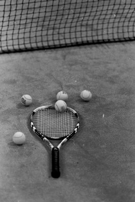 A tennis racket and several tennis balls are arranged on a clay court surface. The net is visible in the background casting a shadow on the court.
