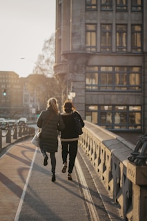 Two individuals walk along a bridge during sunset. They are dressed in warm clothing and are walking closely together, suggesting companionship. The bridge has ornate railings and the background features a large urban building with multiple windows.