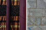 A textured stone wall next to a window with ornate curtains featuring horizontal stripes of orange and black. The wall shows signs of wear and weathering, while the window frame is a deep burgundy color.