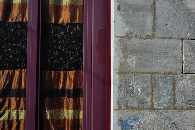 A textured stone wall next to a window with ornate curtains featuring horizontal stripes of orange and black. The wall shows signs of wear and weathering, while the window frame is a deep burgundy color.
