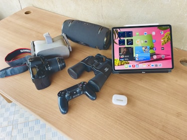 A wooden table displays various electronic devices and tech accessories. There is a tablet with a colorful screen, a digital camera with a strap, virtual reality headset, binoculars, a portable speaker, a game controller, and wireless earbud case.