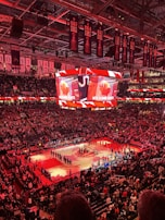 An indoor sports arena filled with spectators and a basketball court illuminated by red lighting. The scoreboards and video screens display promotional content, with numerous banners and championship flags hanging from the ceiling. Players and officials are gathered on the court, possibly in preparation for a game or event.