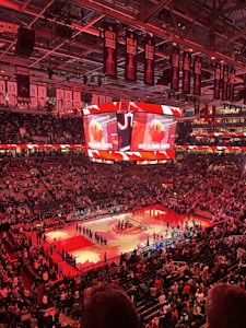 An indoor sports arena filled with spectators and a basketball court illuminated by red lighting. The scoreboards and video screens display promotional content, with numerous banners and championship flags hanging from the ceiling. Players and officials are gathered on the court, possibly in preparation for a game or event.
