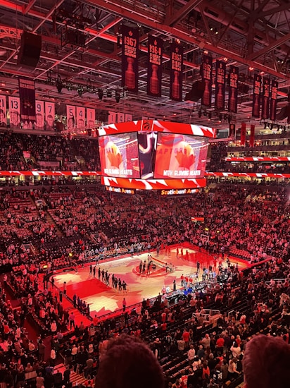 An indoor sports arena filled with spectators and a basketball court illuminated by red lighting. The scoreboards and video screens display promotional content, with numerous banners and championship flags hanging from the ceiling. Players and officials are gathered on the court, possibly in preparation for a game or event.