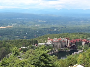 An expansive view of a scenic landscape featuring a large, historic resort complex nestled among lush greenery, with mountains in the background and a serene body of water in the foreground.