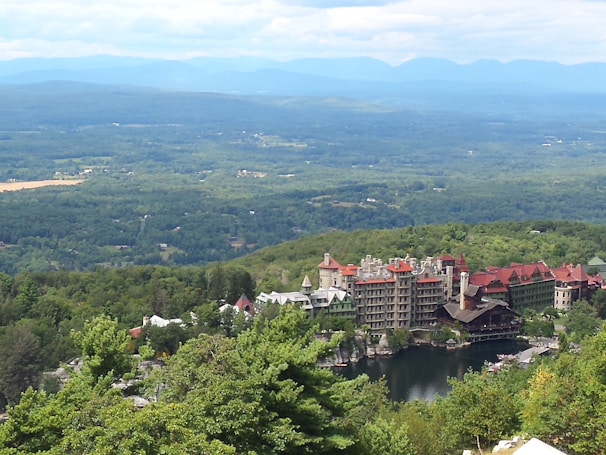 An expansive view of a scenic landscape featuring a large, historic resort complex nestled among lush greenery, with mountains in the background and a serene body of water in the foreground.